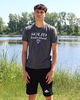 Person wearing a 'SOLID Individual' t-shirt standing by a lake with trees in the background
