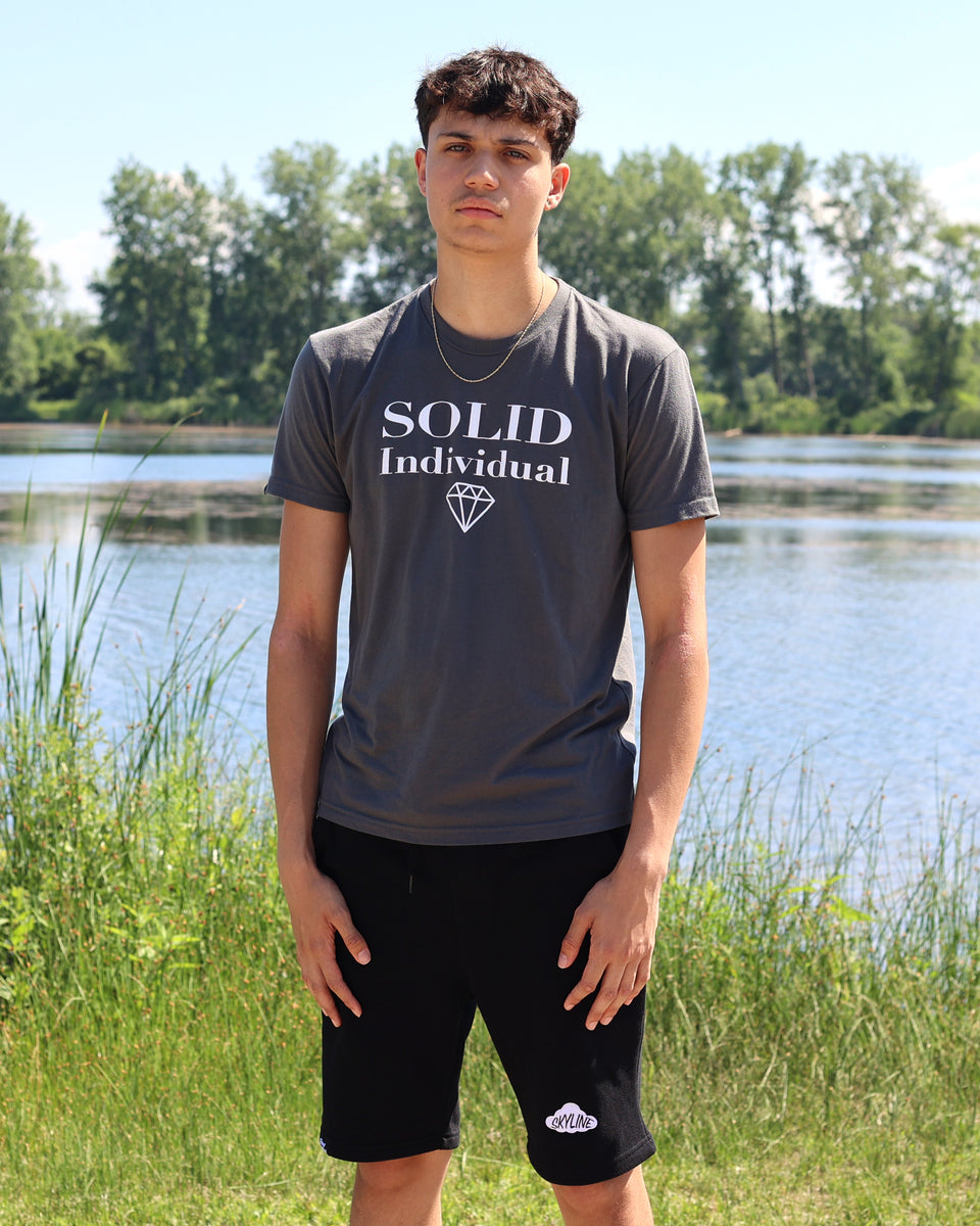 Person wearing a 'SOLID Individual' t-shirt standing by a lake with trees in the background